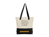 A personalized canvas tote bag with a natural cream upper section, black bottom panel, and black shoulder straps. The front features the text 'ADVOCATE CHRISTIAN ACADEMY' in black with gold accents, and the name 'SAMANTHA' printed in gold on the black bottom section.