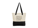 A canvas tote bag with a cream upper section, black bottom panel, and black shoulder straps. The front features the text 'ADVOCATE CHRISTIAN ACADEMY' in black with gold accents.