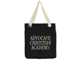 A black canvas tote bag with cream-colored shoulder straps and metal grommets, featuring the text 'ADVOCATE CHRISTIAN ACADEMY' printed in white with gold accents on the front.
