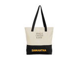 A personalized cotton twill tote bag with a cream upper section, black bottom panel, and black shoulder straps. The front features a 5"x3.125" ADVOCATE CHRISTIAN ACADEMY logo in gold and black lettering, with the name "Samantha" in gold all caps in the black bottom panel.